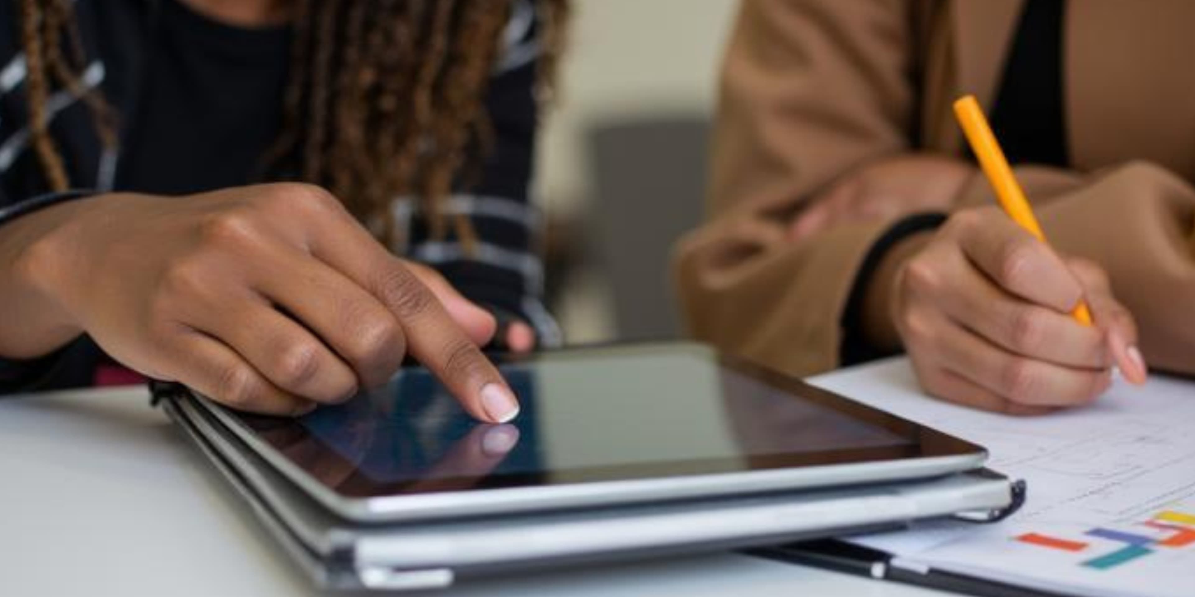 Close-up of hands using a tablet and writing with a pencil on a white desk.